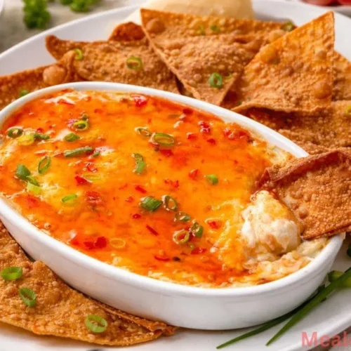 golden-brown baked crab rangoon dip in a white ceramic dish topped with sweet chili sauce and sliced green onions, served with crispy wonton chips